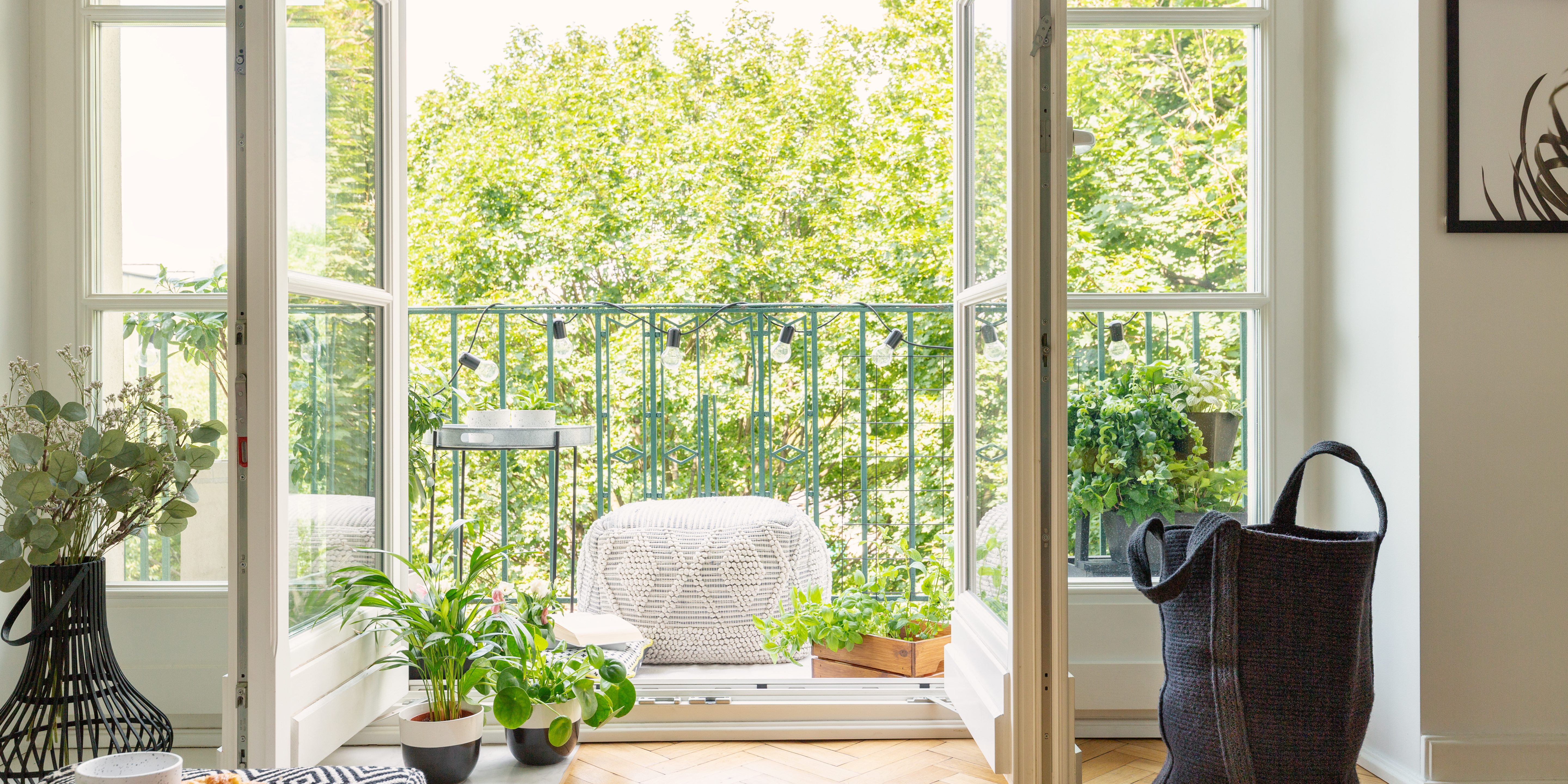 Open glass door from a living room interior into a city garden on a sunny balcony with green plants and comfy furniture